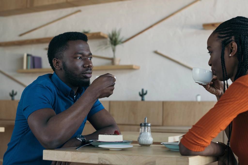 Young Black man and woman sitting down at a table drinking coffee and talking to each other.