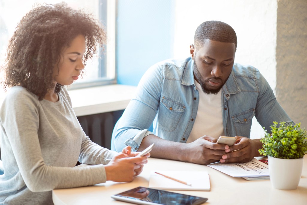 Black man and mixed woman sitting at a table together looking their cell phones.