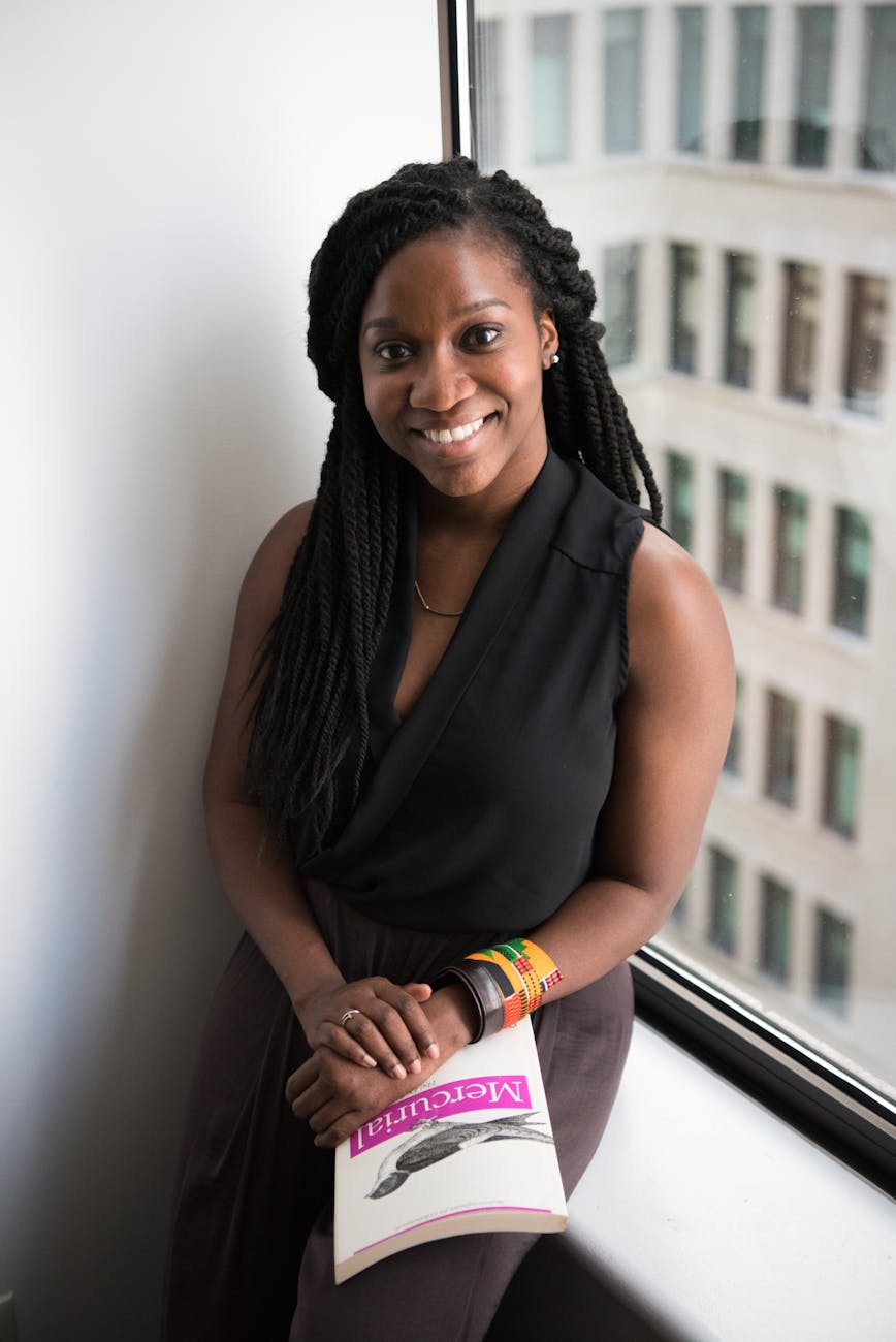 Black young woman with braids smiling at the camera with a book in her lap.