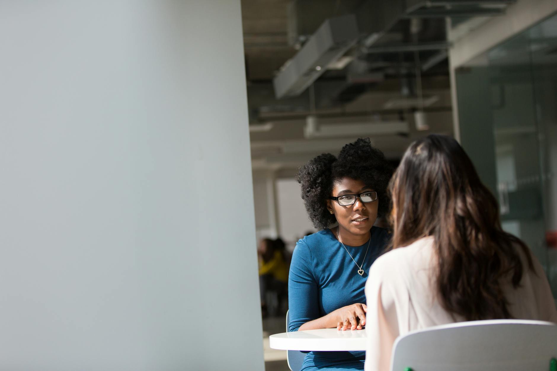 Black woman with afro sitting at table taking to a white woman with brown hair.