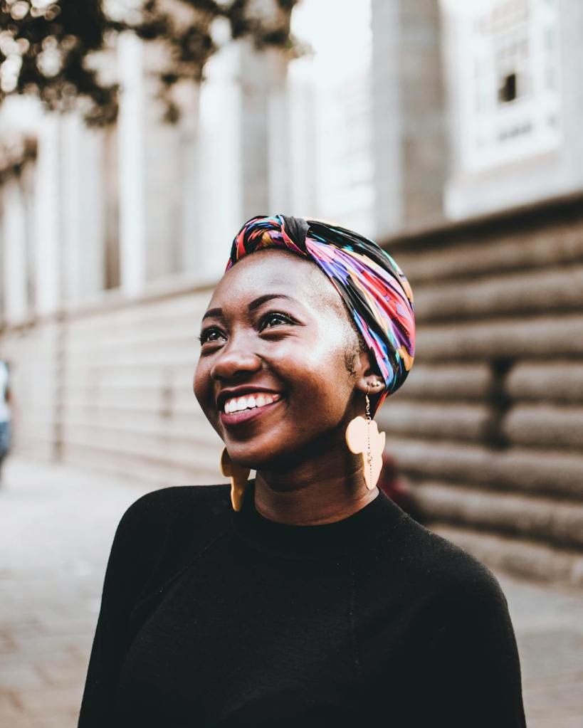 young black woman smiling with color head wrap and gold Africa-shaped earrlings