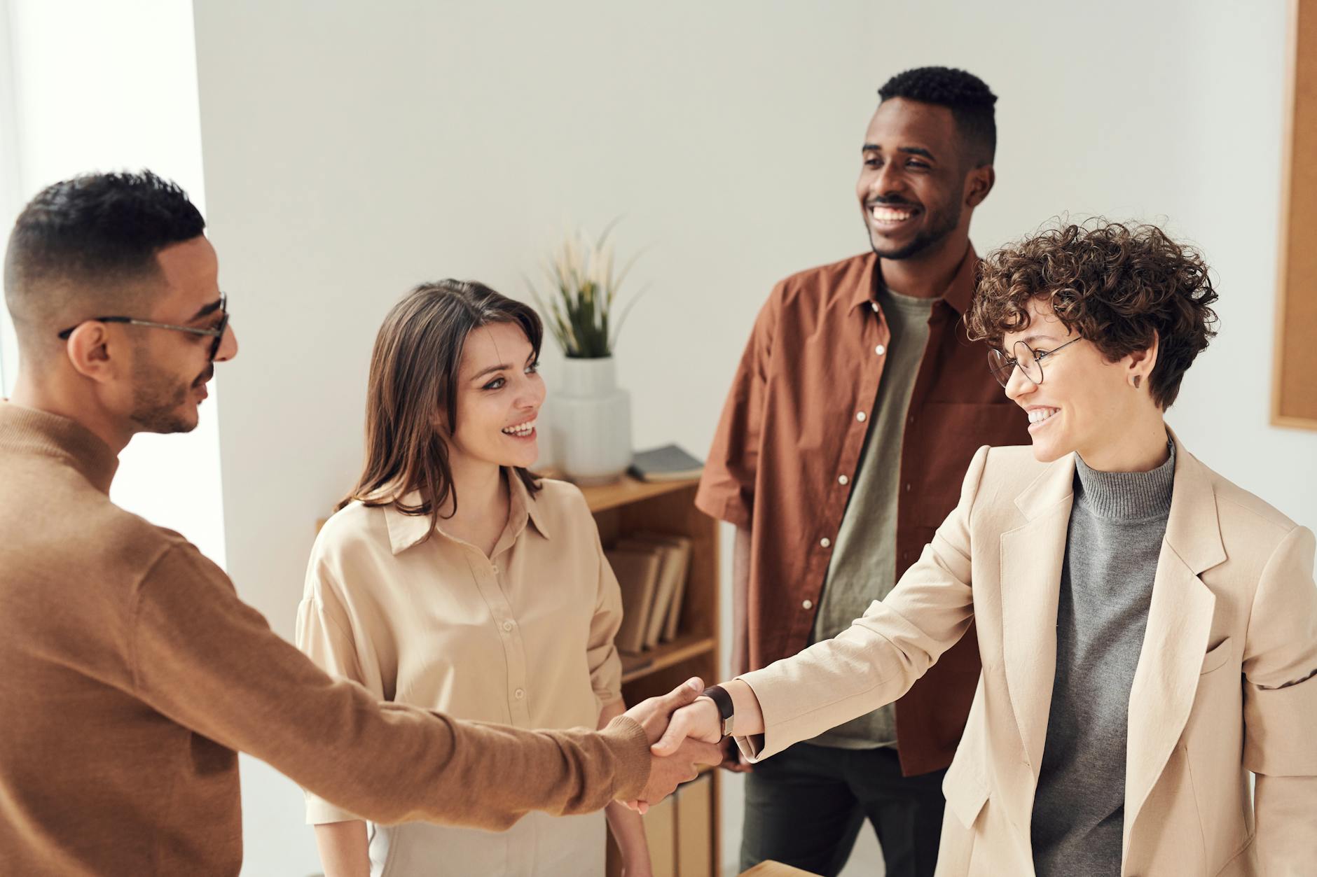 Group of 4 people smiling at each other. Latina woman with curly hair and glasses wearing a beige blazer shakes hands with man wearing glasses and brown sweater.
