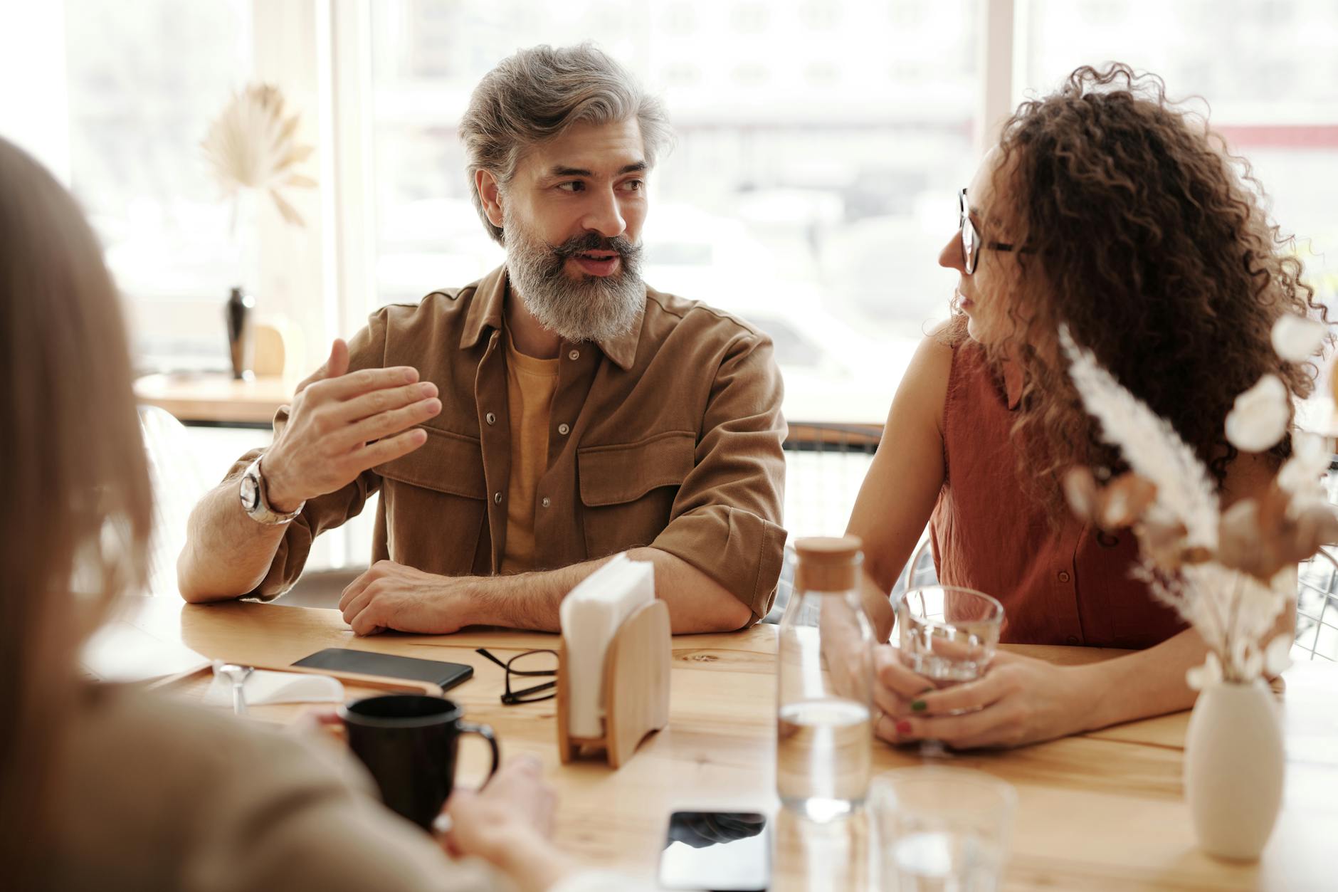 Mature man with thick hair and beard sitting at the table talking to a woman with curly hair and glasses.