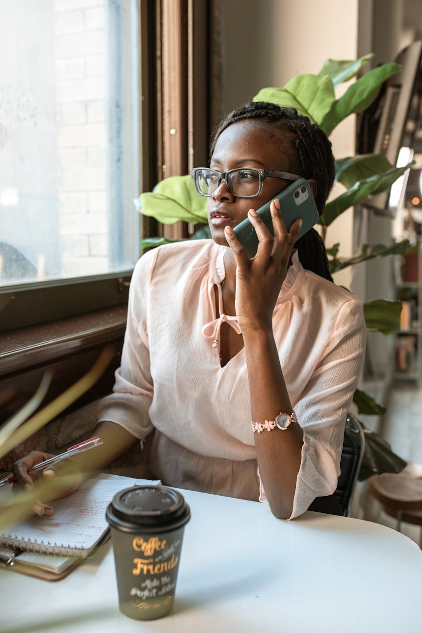Young Black woman sitting at a cafe talking on her cell phone looking out the window with coffee and writing tablets on the table in front of her.