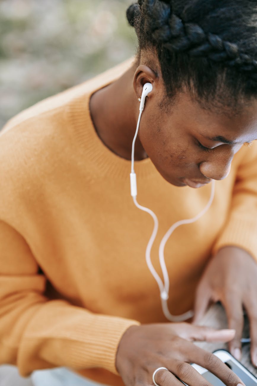 Black woman wearing yellow sweater wearing white earbuds using her cell phone.