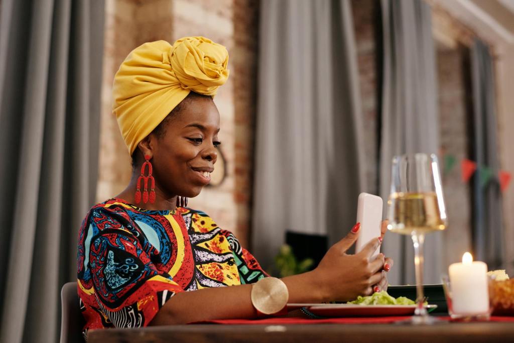 Black woman with yellow head wrap and red long earrings and a colorful top sitting down to dinner with wine and salad smiling at her cell phone.