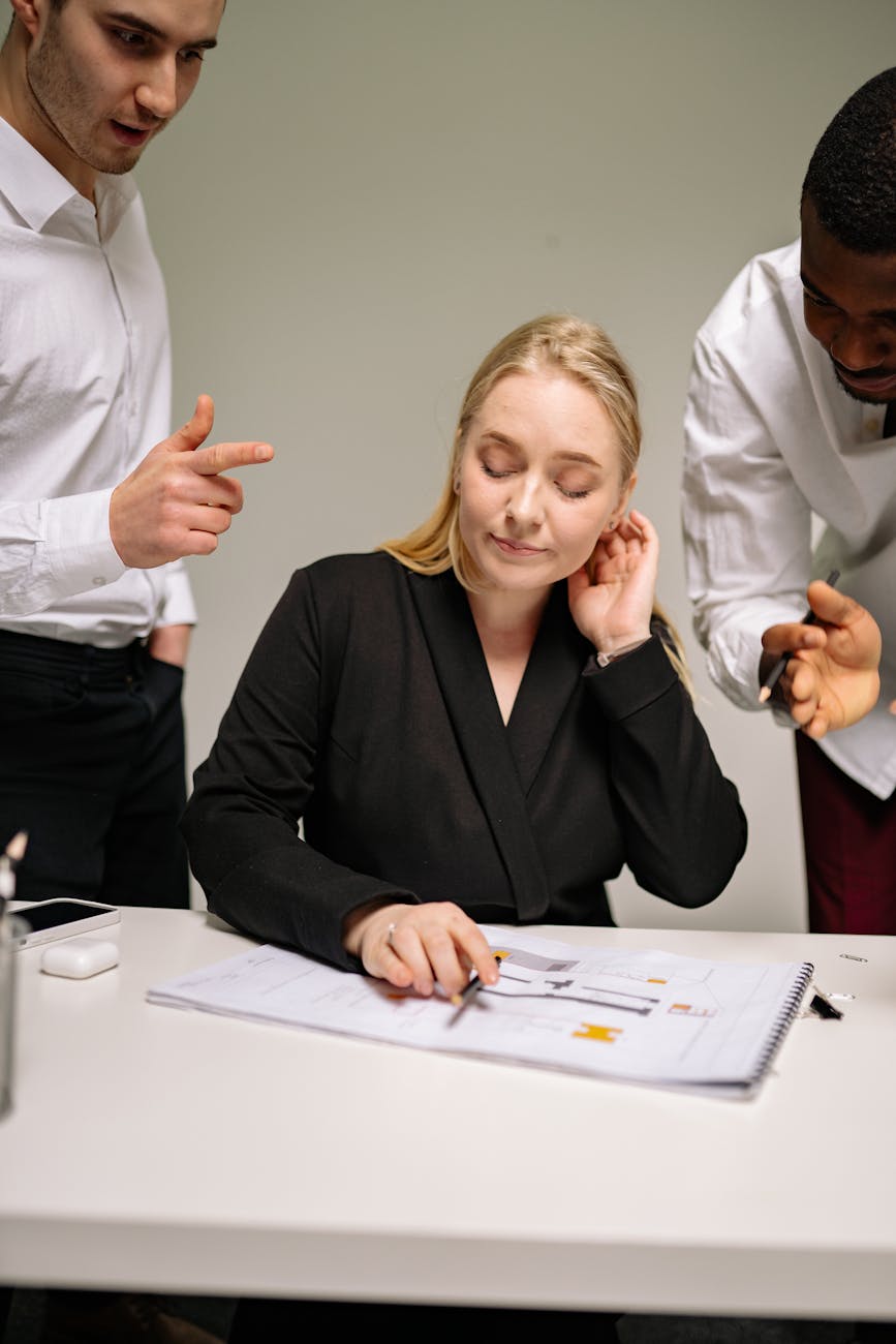 A blonde young woman sitting at a desk looking upset while talking to a white man and Black man.