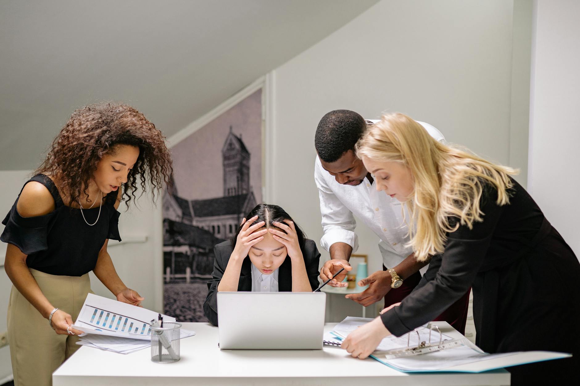 Group of colleagues standing around a desk while woman is sitting down who is grabbing her head looking annoyed.