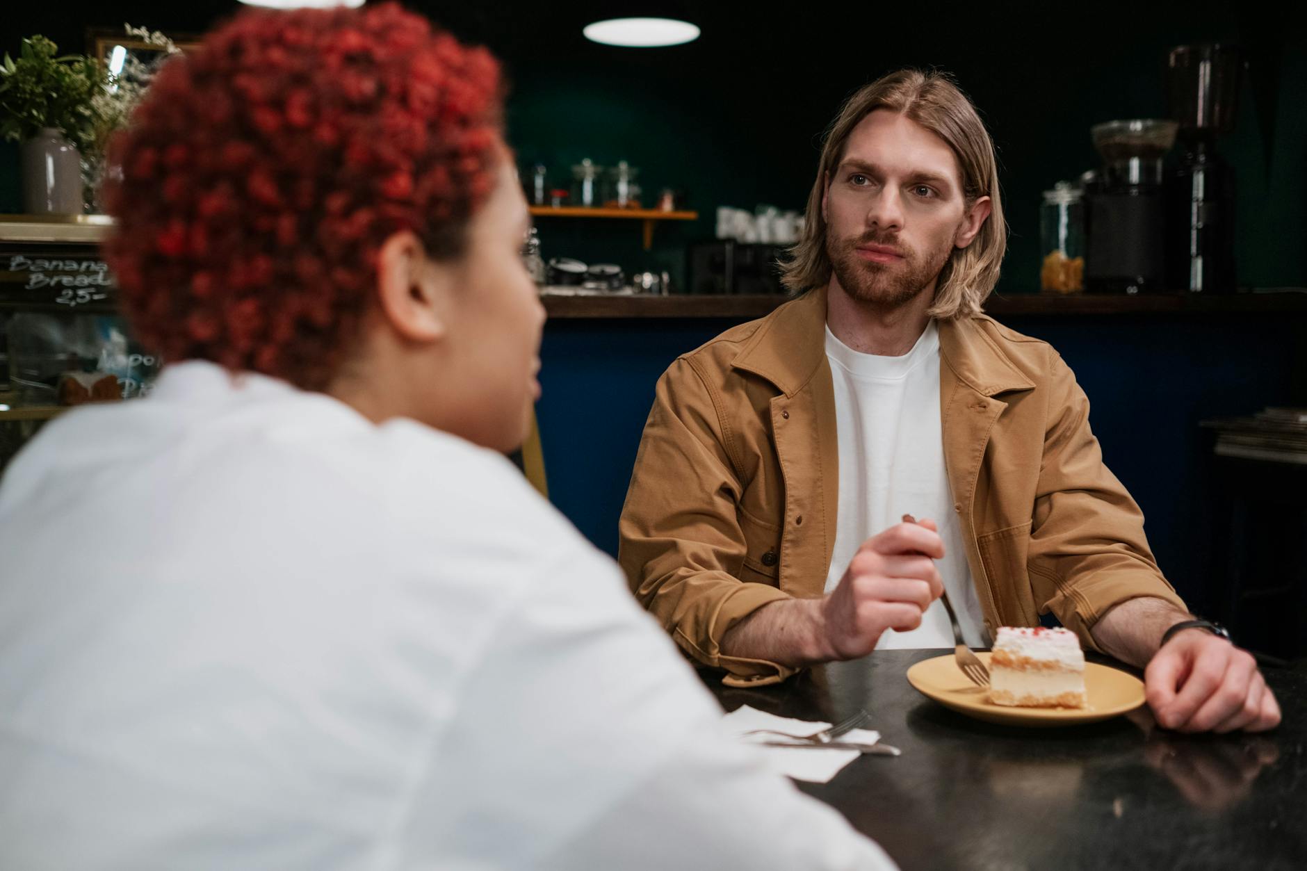 Black woman and White man sit at a table talking while man eats dessert.