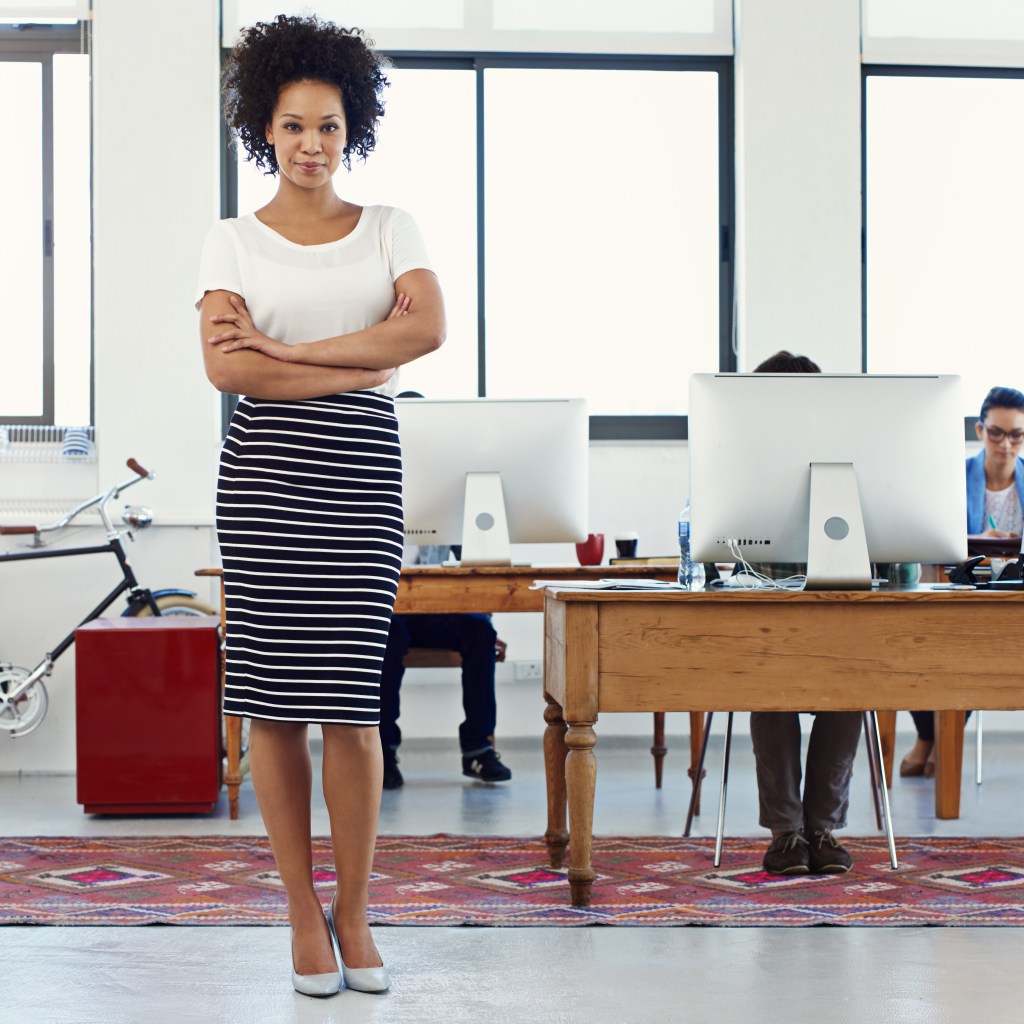 Black woman in skirt standing up in a work office smiling at camera.