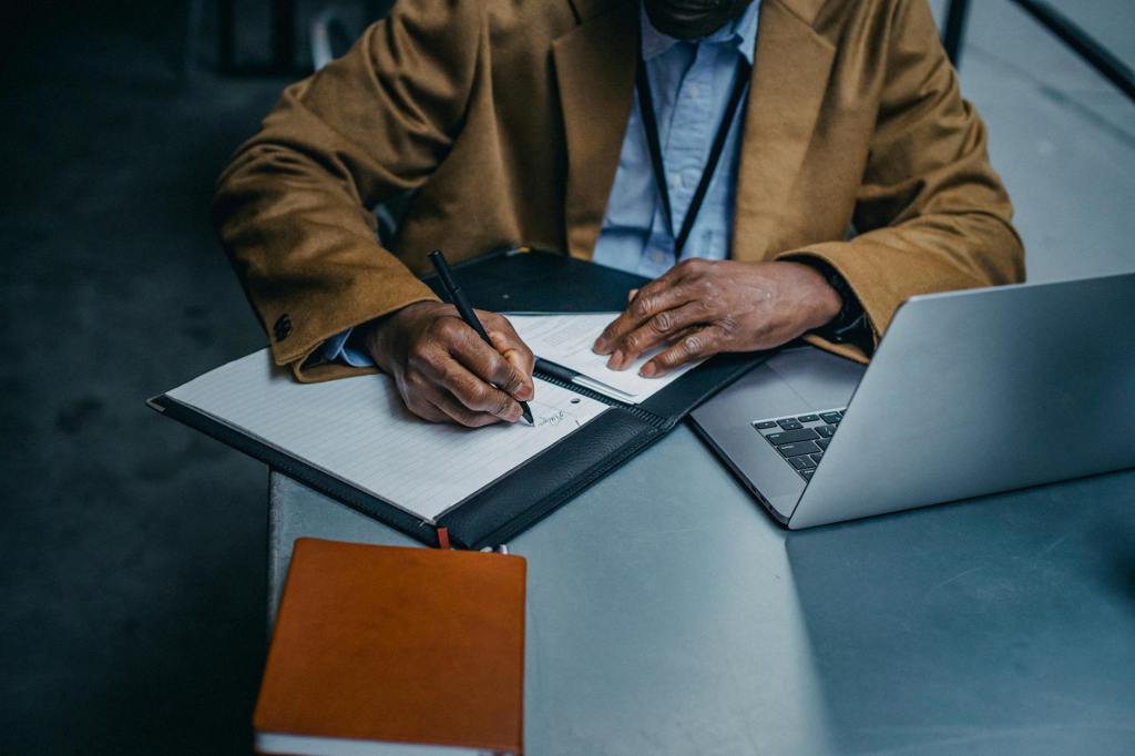 Black person sitting at table with laptop and notebook.