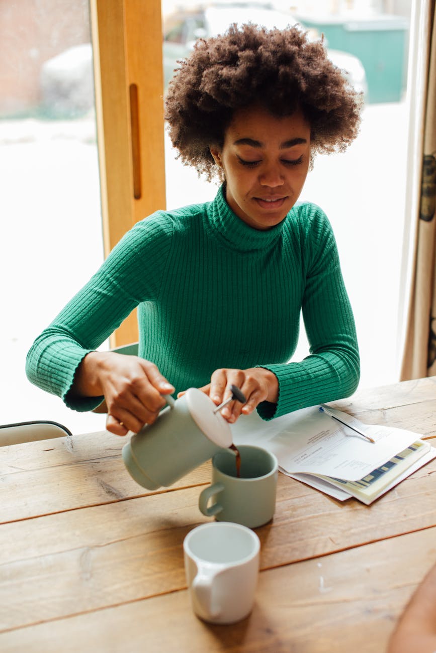 Black woman sitting at a table pouring a cup of coffee.